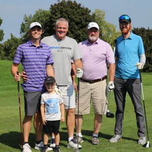 Four men with golf clubs and one child pose smiling at camera on golf course