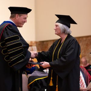 President in graduation regalia shakes hand of women with gray hair graduating