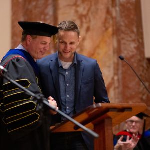 President in graduation regalia shakes hand of man receiving award