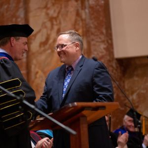 President in graduation regalia shakes hand of man receiving award