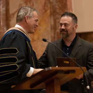 President in graduation regalia shakes hand of man receiving award